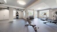 This fitness facility at 6020–6040 Linley Valley Drive in Nanaimo, British Columbia, offers a generously proportioned single-floor exercise room designed to support a full range of workout routines. The space features a clean, modern aesthetic with neutral grey walls, white structural columns, and coffered ceiling details that lend a polished, organized feel throughout. Durable rubber flooring covers the entire area, providing both safety and comfort underfoot.

Natural light flows in through multiple large windows along the perimeter walls, offering pleasant outdoor views of the surrounding landscaping and community. Flush-mount circular ceiling lights supplement the natural light, ensuring the space remains bright and well-lit at all hours.

The facility is comprehensively equipped for both cardio and strength training. Cardio equipment includes treadmills, an elliptical trainer, a stationary bike, and a rowing machine. For strength training, the room features a multi-station cable machine, a full rack of dumbbells, adjustable weight benches, and medicine and stability balls. A wall of mirrors in one section enhances the sense of space and supports form-checking during workouts. Additional amenities include a water fountain and coat/bag rack hooks, adding practical convenience. The layout is open and thoughtfully arranged, allowing multiple users to exercise simultaneously with ample room to move freely.