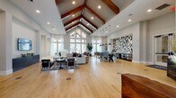 This impressive single-floor communal space is defined by its dramatic vaulted cathedral ceiling adorned with rich, dark-stained exposed wooden beams, creating an immediate sense of grandeur upon entry. Light hardwood flooring spans the entire space, lending warmth and continuity throughout. The welcoming entryway features soaring walls, large transom windows, and curated artwork, setting an inviting tone from the moment you arrive.

The main open-plan area flows seamlessly between distinct functional zones. A comfortable lounge area is anchored by a shiplap accent wall with a mounted flat-screen television, complemented by modern furnishings arranged over a patterned area rug. Adjacent to this is a casual seating nook with leather armchairs and floor-to-ceiling windows that flood the space with natural light and offer views of the surrounding exterior.

A dining and social area features a sturdy wood communal table with woven rope-style chairs, set against a striking geometric tile feature wall in black, white, and grey tones. Pendant lights with warm metallic interiors hang above, adding ambient sophistication. A kitchenette with countertop appliances and cabinetry backs this dining zone seamlessly.

Private rooms accessible via glass-paneled doors provide additional flexibility, while the overall palette of soft grey walls, natural wood tones, and contemporary furnishings creates a cohesive, modern atmosphere throughout.