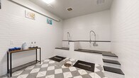 Located within the Westhouse community at 11325 Park Vista Blvd in Fort Worth, Texas, this purpose-built pet spa offers a thoughtfully designed grooming space for residents and their pets. The room features white subway tile walls trimmed with dark accent borders, creating a clean and polished aesthetic throughout. The bold black-and-white geometric patterned tile flooring adds a stylish, contemporary touch while remaining practical and easy to maintain. Two elevated wash stations with stainless steel sprayer fixtures are positioned along one wall, each accessible via built-in steps — accommodating pets of all sizes with ease. The floor-level drains ensure efficient water management during wash sessions. A slim console table along the opposite wall holds grooming supplies, including shampoo dispensers and accessories, keeping everything conveniently within reach. Whimsical pet-themed artwork displayed on the walls adds a welcoming, cheerful ambiance. The space is climate-controlled with a wall-mounted AC unit and fan, ensuring comfort year-round. A glass-panel entry door and interior window wall provide visibility into the adjoining area, while overhead lighting keeps the room bright and functional. This well-appointed pet spa delivers a practical yet stylish environment for pet grooming.