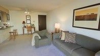 This single-floor residential space offers a warm and functional layout designed for comfortable everyday living. The living area serves as the heart of the home, featuring plush wall-to-wall carpeting in a soft neutral tone that adds a sense of warmth underfoot. The room is furnished with a cushioned sofa and matching armchair in muted sage green, accented with decorative pillows in earth tones, creating an inviting and cohesive aesthetic. A ceiling fan with integrated lighting provides both comfort and illumination, while a table lamp adds a softer, ambient glow to the space. The living area also accommodates a compact desk and chair tucked neatly along one wall, offering a practical workspace alongside framed artwork that adds a personal touch. The smooth, light-colored walls reflect natural and artificial light evenly throughout the room, contributing to a bright and open feel. A dark wood entry door provides a clean visual anchor to the space. Overall, the interior presents a practical and comfortable environment with a calm, neutral palette that makes it easy to personalize and settle into.