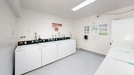 Located at 14300 Mulberry Dr in Whittier, California, this shared laundry facility offers a clean, efficiently organized space designed for practical, everyday use. The room is finished with bright white walls and light speckled flooring, creating a clean and functional atmosphere throughout.

A row of top-loading washing machines lines one wall, each equipped with modern digital controls and WASH-branded card payment systems, allowing for convenient cashless transactions. Opposite the washers, a lineup of front-loading dryers provides ample drying capacity. A long, flat surface above the dryers offers useful space for sorting and folding laundry.

The facility is well-lit by overhead fluorescent lighting, supplemented by natural light from louvered ventilation windows positioned near the ceiling. A skylight further brightens the interior, contributing to the open, airy feel of the room. Instructional and usage signage is clearly posted on the walls, including laundry system guides and facility rules, ensuring users can operate the machines with ease.

Additional amenities include a payment kiosk station mounted on the wall and a security camera for resident safety. The room opens to an exterior entryway through a glass-panel door, providing convenient access. Overall, the facility is well-maintained and thoughtfully equipped to serve the laundry needs of the surrounding residential community.