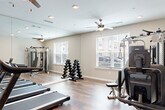 Located at 6565 Chupp Rd in Stonecrest, GA, this fitness center is a well-appointed single-room facility designed to support a comprehensive workout routine. The space features rich, hardwood-style flooring throughout, lending a clean and polished aesthetic that complements the neutral-toned walls and bright white trim.

Cardio equipment is a focal point of the room, with multiple Spirit treadmills arranged in a row alongside stationary and recumbent bikes. For strength training, a multi-functional cable machine with an adjustable bench provides versatile resistance options, while a full dumbbell rack offers a range of free weights for targeted exercises. Floor-to-ceiling mirrors line one wall, visually expanding the space and allowing users to monitor their form during workouts.

Natural light flows in through multiple windows fitted with white blinds, creating a bright and energizing atmosphere. Ceiling fans with integrated lighting are positioned throughout the room to maintain comfortable airflow. Recessed lighting supplements the natural light, ensuring the space remains well-illuminated at all times. A wall-mounted flat-screen television is positioned near the treadmills, providing entertainment during cardio sessions. The overall ambiance is clean, functional, and welcoming, making this fitness center a practical and inviting space for daily exercise.