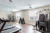 Located at 6565 Chupp Rd in Stonecrest, GA, this fitness center is a well-appointed single-room facility designed to support a comprehensive workout routine. The space features rich, hardwood-style flooring throughout, lending a clean and polished aesthetic that complements the neutral-toned walls and bright white trim.

Cardio equipment is a focal point of the room, with multiple Spirit treadmills arranged in a row alongside stationary and recumbent bikes. For strength training, a multi-functional cable machine with an adjustable bench provides versatile resistance options, while a full dumbbell rack offers a range of free weights for targeted exercises. Floor-to-ceiling mirrors line one wall, visually expanding the space and allowing users to monitor their form during workouts.

Natural light flows in through multiple windows fitted with white blinds, creating a bright and energizing atmosphere. Ceiling fans with integrated lighting are positioned throughout the room to maintain comfortable airflow. Recessed lighting supplements the natural light, ensuring the space remains well-illuminated at all times. A wall-mounted flat-screen television is positioned near the treadmills, providing entertainment during cardio sessions. The overall ambiance is clean, functional, and welcoming, making this fitness center a practical and inviting space for daily exercise.