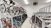 This single-level indoor bicycle storage room is purpose-built to accommodate a large number of bikes in an organized and secure environment. The space is defined by its galvanized steel double-decker bicycle racks, which run along multiple walls and through the center of the room, maximizing vertical storage capacity. The lower tier allows for easy roll-in access, while the upper tier utilizes a ramp-style rail system to slide bikes into elevated positions, effectively doubling the number of bikes that can be stored within the footprint.

The room features clean white walls and a light-toned hard flooring surface that is easy to maintain. Overhead, flush-mounted fluorescent light fixtures provide consistent, bright illumination throughout the space. A ceiling-mounted heater ensures the room remains climate-controlled, making it practical for year-round use in colder climates.

Access is managed through a secure door equipped with an electronic key-fob lock, providing controlled entry. A floor pump is available on-site, adding a practical convenience for cyclists. The 3D floorplan reveals a well-configured layout with clear pathways between rack rows, allowing users to navigate and retrieve bikes with ease. Overall, the space is functional, well-lit, and designed with high-capacity, organized bicycle storage as its primary purpose.