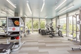 This fitness center is thoughtfully designed to support a comprehensive workout experience across multiple distinct zones. The cardio area is defined by floor-to-ceiling windows that flood the space with natural light and frame views of the surrounding greenery and streetscape, creating an energizing atmosphere. Equipment here includes treadmills, elliptical machines, and a rowing machine, all positioned to take advantage of the outdoor views. The flooring in this zone features light-toned, patterned carpet tiles that complement the bright, teal-painted ceiling — a standout design element that lends the space a modern, uplifting character. Linear LED lighting runs throughout, providing consistent, clean illumination. A separate strength and free weights area transitions into the space, featuring wood-look flooring, weight benches, a dumbbell rack, and an accent wall with a decorative geometric pattern in neutral tones with colorful insets. A dedicated cycling studio with spin bikes and a large display screen is accessible via a connecting corridor, offering a more immersive group-style workout environment. The overall design is polished and cohesive, combining functional layout with contemporary finishes.