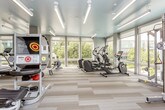 This fitness center is thoughtfully designed to support a comprehensive workout experience across multiple distinct zones. The cardio area is defined by floor-to-ceiling windows that flood the space with natural light and frame views of the surrounding greenery and streetscape, creating an energizing atmosphere. Equipment here includes treadmills, elliptical machines, and a rowing machine, all positioned to take advantage of the outdoor views. The flooring in this zone features light-toned, patterned carpet tiles that complement the bright, teal-painted ceiling — a standout design element that lends the space a modern, uplifting character. Linear LED lighting runs throughout, providing consistent, clean illumination. A separate strength and free weights area transitions into the space, featuring wood-look flooring, weight benches, a dumbbell rack, and an accent wall with a decorative geometric pattern in neutral tones with colorful insets. A dedicated cycling studio with spin bikes and a large display screen is accessible via a connecting corridor, offering a more immersive group-style workout environment. The overall design is polished and cohesive, combining functional layout with contemporary finishes.
